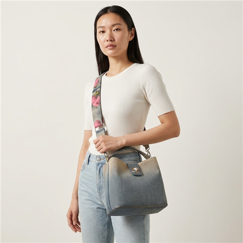 Betsey Johnson Denim Bucket Bag featuring faded blue and neutral ombre coloring, silver tone hardware, short top handle, and a wide adjustable strap with pink and yellow floral details.
