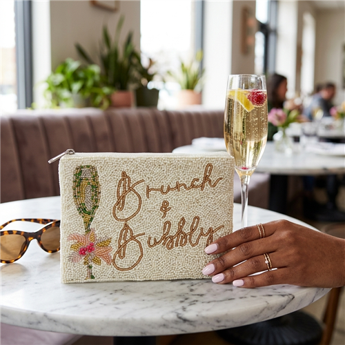 Fashion Culture ivory beaded zip pouch featuring Brunch and Bubbly lettering, a green and gold tone champagne flute, pink floral bow detail, and white zipper.